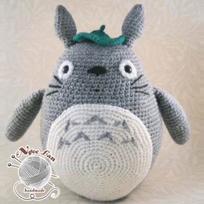 Un peluche tejido a crochet hecho a mano que se asemeja a Totoro, un personaje de "Mi vecino Totoro" de Studio Ghibli. El peluche es gris con vientre blanco, nariz negra y ojos redondos, con una pequeña hoja en la cabeza. En la esquina inferior izquierda se ve un logo con "El Vroc Lana hecho a mano".