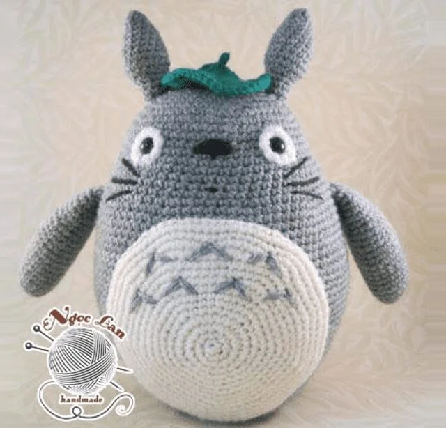 Un Peluche Tejido a Crochet Hecho a Mano Que Se Asemeja a Totoro Un Personaje De mi Vecino Totoro De Studio Ghibli El Peluche Es Gris Con Vientre Blanco Nariz Negra Y Ojos Redondos Con Una Pequeña Hoja En La Cabeza En La Esquina Inferior Izquierda Se Ve Un Logo Con el Vroc Lana Hecho a Mano