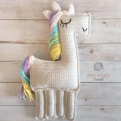 Una llama de ganchillo se encuentra sobre una superficie de madera. La llama es blanca con una melena multicolor, con tonos pastel de rosa, azul, amarillo y verde. Sus ojos están cerrados, representados con simples puntadas de hilo negro. El logotipo "Spin a Yarn Crochet" es visible al lado.