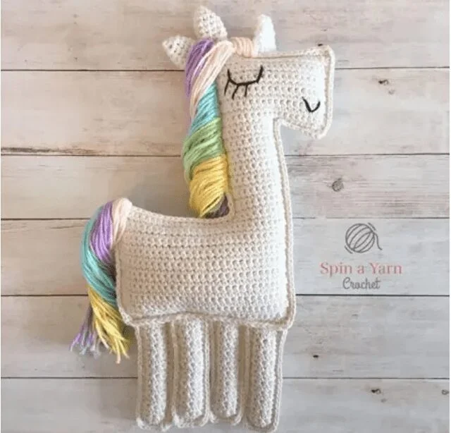 Una Llama De Ganchillo Se Encuentra Sobre Una Superficie De Madera La Llama Es Blanca Con Una Melena Multicolor Con Tonos Pastel De Rosa Azul Amarillo Y Verde Sus Ojos Están Cerrados Representados Con Simples Puntadas De Hilo Negro El Logotipo spin a Yarn Crochet Es Visible Al Lado