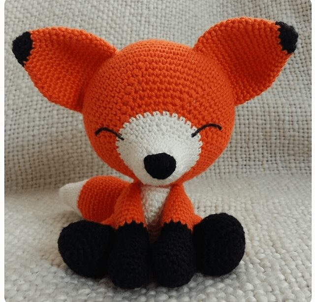 Un Zorro De Peluche De Ganchillo Con Cuerpo Naranja Hocico Vientre Y Punta De Cola Blancos Y Patas Y Nariz Negras Está Sentado Sobre Un Fondo Con Textura Gris El Zorro Parece Sonreír Con Los Ojos Cerrados Mostrando Una Expresión Linda Y Juguetona