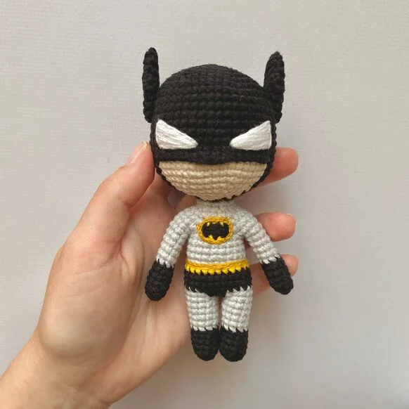 Una Mano Sostiene Un Pequeño Muñeco De Ganchillo Que Se Parece a Batman La Muñeca Tiene Una Máscara Negra Con Orejas Puntiagudas Parches Blancos En Los Ojos Y Una Insignia Negra Y Amarilla En El Pecho Viste Un Traje Gris Con Guantes Y Botas Negros Y Un Cinturón Amarillo