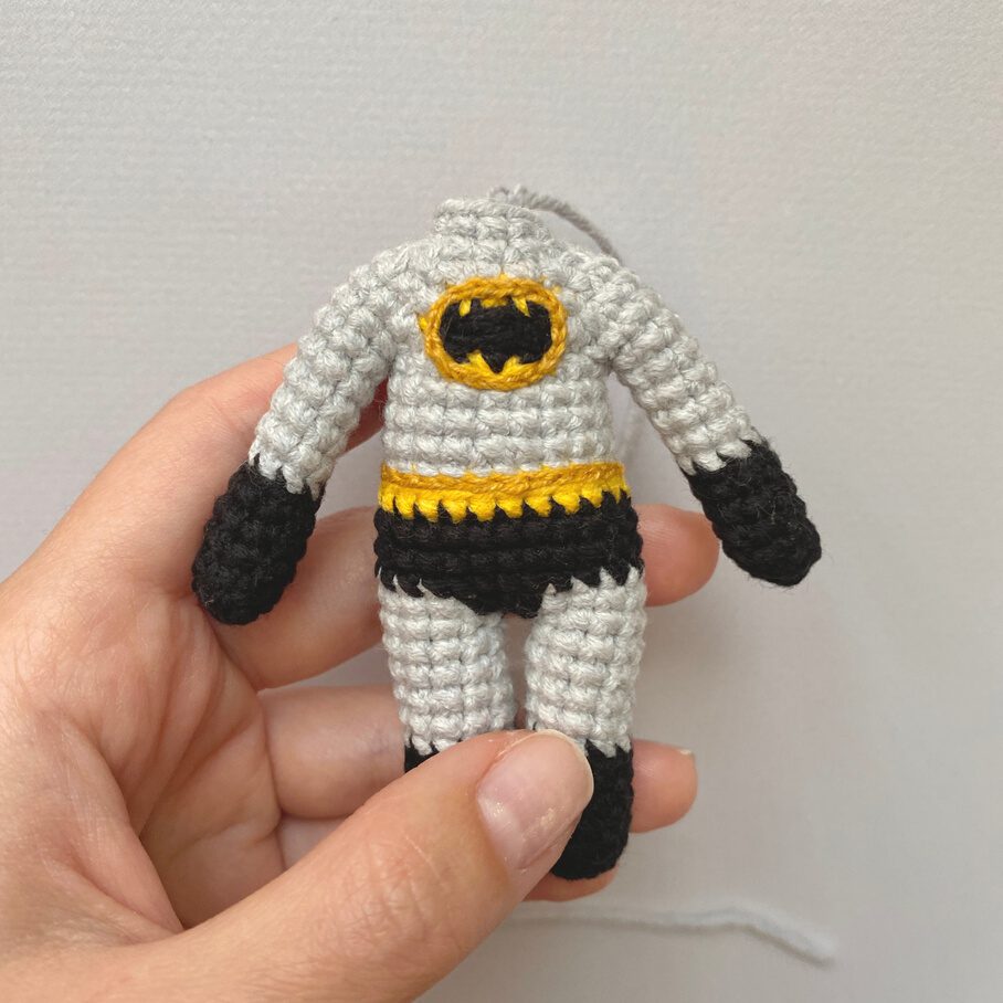 Una Mano Sostiene Una Pequeña Muñeca De Ganchillo Que Se Asemeja a Un Disfraz De Superhéroe Con Un Traje Gris Con El Emblema Amarillo Y Negro De Batman En El Pecho Guantes Negros Y Pantalones Cortos Negros La Muñeca No Tiene Cabeza Ni Extremidades Lo Que Resalta El Detallado Disfraz