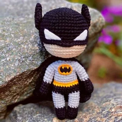 Un pequeño juguete de ganchillo hecho a mano que se parece a Batman está colocado contra una roca. Tiene la icónica máscara, capa y botas negras, un mono gris con un cinturón amarillo con el símbolo del murciélago negro en el centro. El fondo incluye sutil vegetación y flores.