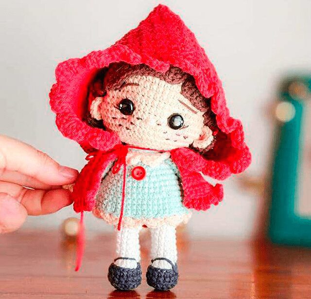 Una Mano Sostiene Una Muñeca Tejida Que Se Parece a Caperucita Vestida Con Una Capa Roja Con Capucha Y Un Pequeño Botón Rojo La Muñeca Tiene Ojos Negros Rubor En Las Mejillas Y Cabello Castaño Corto Está Vestido Con Un Traje Azul Claro Y Zapatos Blancos El Fondo Está Borroso Y Muestra Objetos Confusos