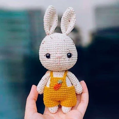 Un conejo de juguete tejido a crochet con grandes ojos negros está erguido. Viste un overol amarillo adornado con un pequeño detalle de zanahoria en el frente. La mano de una persona sostiene el juguete, con un fondo borroso visible detrás.