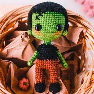 Un lindo muñeco de crochet que se parece a Frankenstein está sentado en una canasta de mimbre. La muñeca tiene piel verde brillante, grandes ojos negros, cabello negro y está vestida con una sencilla camisa negra y pantalones de color marrón rojizo. La cesta contiene papel marrón arrugado.