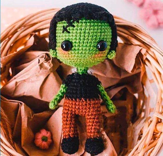 Un Lindo Muñeco De Crochet Que Se Parece a Frankenstein Está Sentado En Una Canasta De Mimbre La Muñeca Tiene Piel Verde Brillante Grandes Ojos Negros Cabello Negro Y Está Vestida Con Una Sencilla Camisa Negra Y Pantalones De Color Marrón Rojizo La Cesta Contiene Papel Marrón Arrugado