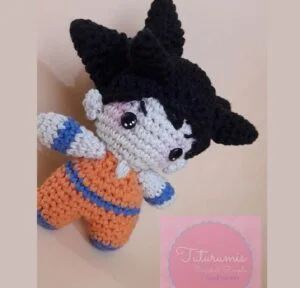 Un muñeco de ganchillo que se parece a Goku con cabello negro puntiagudo, grandes ojos redondos y vestido con un traje naranja y azul. La imagen incluye una etiqueta rosa en la parte inferior con texto en cursiva que dice "Titirumis Crochet Simple".