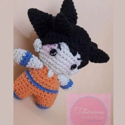 Un muñeco de ganchillo que se parece a Goku con cabello negro puntiagudo, grandes ojos redondos y vestido con un traje naranja y azul. La imagen incluye una etiqueta rosa en la parte inferior con texto en cursiva que dice "Titirumis Crochet Simple".