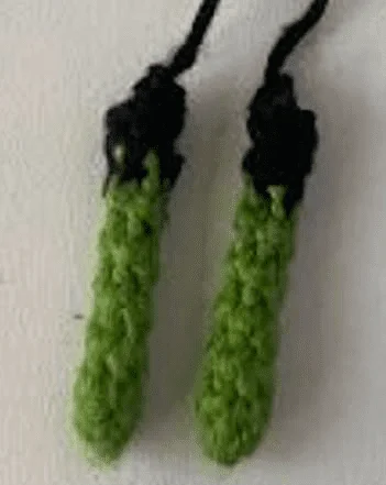 Dos Tiras De Chile Verde Tejidas a Crochet Con Tapas Negras Colgando Una Al Lado De La Otra Que Recuerdan La Estética De Frankenstein Los Chiles Tienen Una Apariencia Texturizada Y Están Colocados Sobre Un Fondo Blanquecino