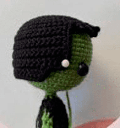 Un Muñeco De Crochet Con Cara Verde Y Pelo Negro Que Recuerda a Frankenstein Que Lleva Una Pequeña Cuenta Blanca a Modo De Ojo La Muñeca Está Vestida Con Un Traje Negro Y Está Colocada Sobre Un Fondo De Color Claro Como Se Muestra En Una Vista Lateral