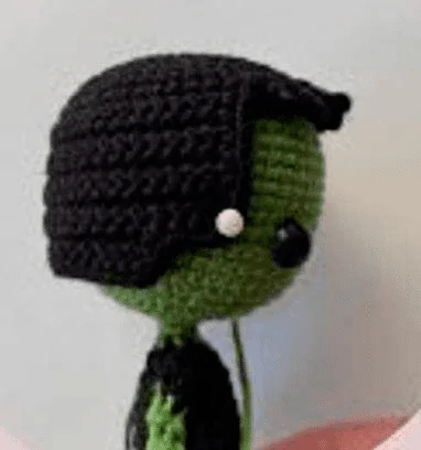 Un Muñeco De Crochet Con Cara Verde Y Pelo Negro Que Recuerda a Frankenstein Que Lleva Una Pequeña Cuenta Blanca a Modo De Ojo La Muñeca Está Vestida Con Un Traje Negro Y Está Colocada Sobre Un Fondo De Color Claro Como Se Muestra En Una Vista Lateral