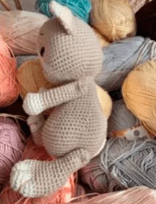 Un Gatito De Peluche Gris Tejido a Crochet Está Rodeado De Coloridas Bolas De Hilo En Tonos De Rosa Amarillo Azul Y Crema El Gato Está Colocado De Lado Y Se Ven Sus Costuras Detalladas