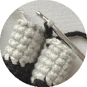 Primer Plano De Un Proyecto De Crochet Que Presenta Dos Piezas Cilíndricas De Crochet En Hilo Gris Claro Junto a Un Ganchillo Se Intercala Hilo Negro Insinuando Un Detalle De Puntada O Patrón Inspirado En Batman El Fondo Es Una Superficie De Tela Neutra