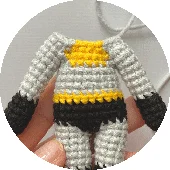 Un Primer Plano De Una Mano Que Sostiene Una Figura De Batman Tejida a Crochet Sin Cabeza La Figura Hecha a Mano Está Vestida Con Un Traje Colorido De Hilo Gris Negro Y Amarillo El Fondo Está Desenfocado Y Se Puede Ver Algo De Hilo Blanco En La Esquina Superior