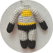 Un Pequeño Muñeco De Crochet Hecho a Mano Inspirado En Batman Con Cuerpo Gris Manos Y Pies Negros Y Un Estampado Amarillo En El Pecho La Muñeca Carece De Rasgos Faciales Y Cuelga Sobre Un Fondo Neutro