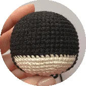 Una Mano Que Sostiene Un Objeto Pequeño Y Redondo De Crochet Con La Parte Superior Negra Y La Parte Inferior Beige Que Se Asemeja a Una Cabeza Simplificada Con Cabello El Diseño Alude Sutilmente a La Icónica Capucha De Batman El Fondo Es Sencillo Y Desenfocado