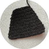 Una Imagen En Primer Plano De Una Pieza De Crochet Negra De Forma Triangular Con Un Patrón Texturizado Evoca La Silueta Icónica De La Capa De Batman La Pieza Está Colocada Sobre Un Fondo Gris Claro Ligeramente Texturizado Y Un Hilo Suelto Se Extiende Desde La Parte Superior Del Trabajo De Crochet