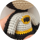 Primer Plano De Una Figura Tejida a Crochet Vestida Con Un Disfraz Inspirado En Batman Con Un Top Gris Con Un Símbolo De Murciélago Negro Y Amarillo En El Pecho Se Ven El Brazo De La Figura Y Parte De La Cabeza Adornada Con Diminutas Orejas De Batman Que Está Sostenida Por Una Mano