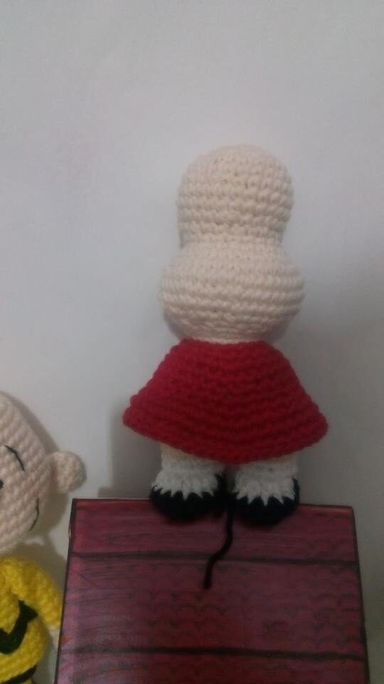 Un Muñeco De Crochet Mirando Hacia Atrás Con La Cabeza El Torso Y Las Piernas Visibles La Muñeca Tiene La Cabeza Blanca Piernas Blancas Con Zapatos Negros Y Un Vestido Rojo Que Recuerda a Mafalda Otra Figura De Crochet Parcialmente Visible Se Encuentra a La Izquierda