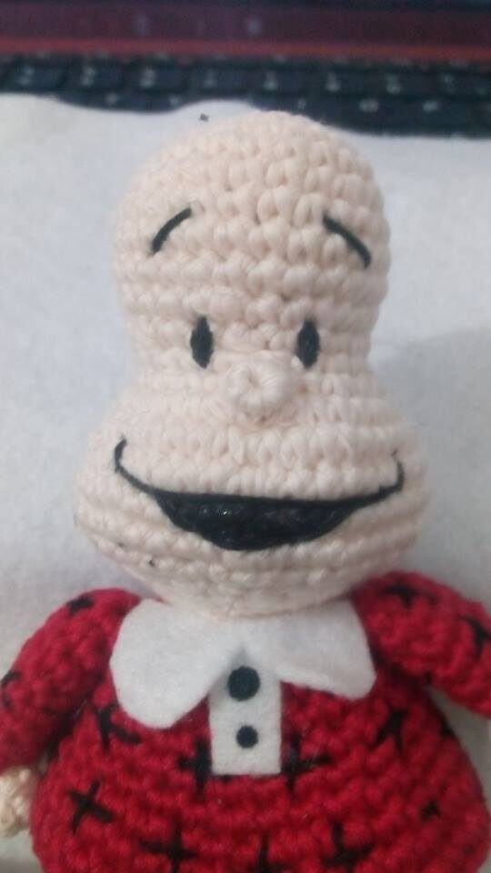 Muñeco De Crochet Con Una Cabeza Grande De Forma Ovalada Y Una Boca Ancha Y Sonriente La Muñeca Tiene Ojos Pequeños Y Negros Y Algunas Líneas En La Frente Viste Una Camisa Roja Con Dibujos Entrecruzados Negros Y Un Cuello Blanco Con Botones Negros Que Recuerda a Mafalda Recostada Sobre Una Superficie Blanca