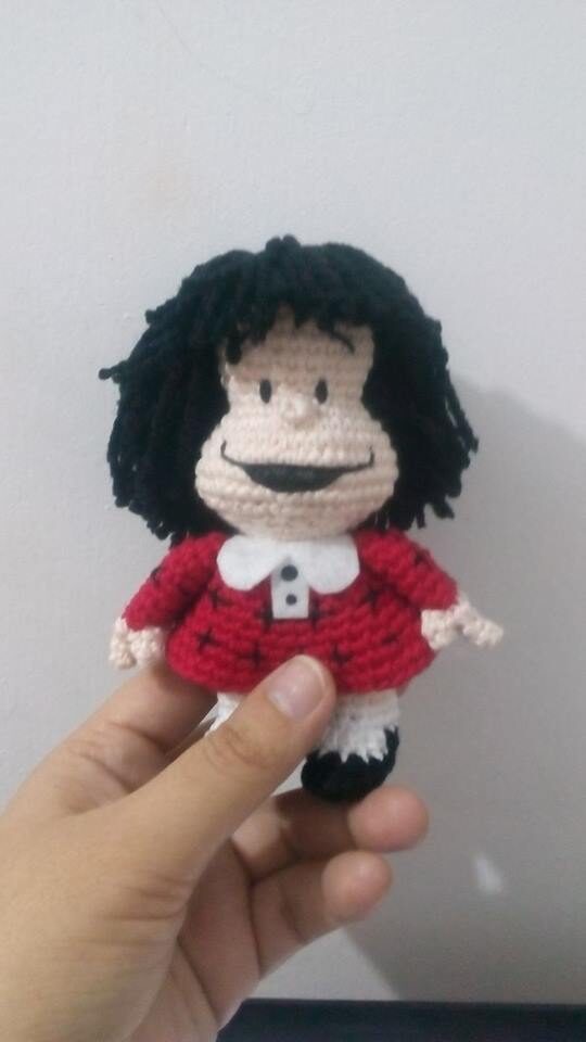 Una Mano Sostiene Una Pequeña Muñeca Mafalda De Crochet Con El Pelo De Hilo Negro La Muñeca Lleva Un Vestido Rojo Con Cuello Blanco Y Zapatos Negros El Fondo Es Una Simple Pared Blanca