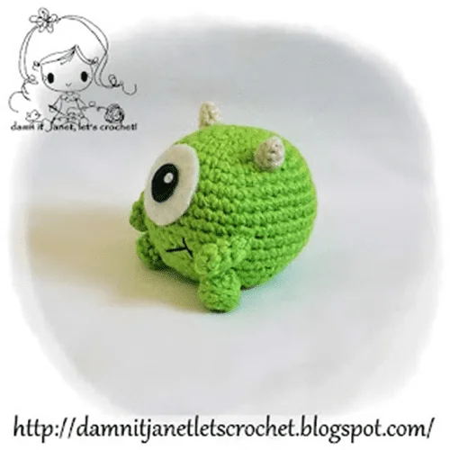 Un Pequeño Monstruo Verde Tejido a Crochet Que Se Parece a Mike Wazowski Con Un Ojo Grande Dos Cuernos Diminutos Y Tres Patas Sentado Sobre Una Superficie Blanca La Imagen Presenta Un Logotipo En La Esquina Superior Izquierda Con Una Niña De Dibujos Animados Y El Texto maldita Sea Janet Vamos a Tejer Una Url En La Parte Inferior Dice httpdamnitjanetletscrochetblogspotcom