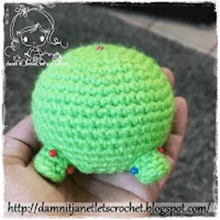 Una Mano Sostiene Una Pequeña Figura De Amigurumi De Crochet Verde Que Se Parece a Mike Wazowsky Con Un Cuerpo Redondo Y Extremidades Diminutas La Figura Tiene Alfileres Rojos Y Azules Incrustados El Fondo Presenta Un Suelo Alfombrado El Texto En La Parte Inferior Dice httpdamnitjanetletscrochetblogspotcom