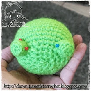 Una Mano Sostiene Un Objeto Pequeño Y Redondo Tejido a Crochet Con Hilo Verde Brillante Que Se Parece a Mike Wazowski El Objeto Tiene Dos Pequeñas Protuberancias En Un Lado Cada Una Con Un Alfiler De Colores Insertado En La Esquina Superior Izquierda Hay Una Marca De Agua Con Una Caricatura Se Muestra Una Url En La Parte Inferior Httpdamnitjanetletscrochetblogspotcom