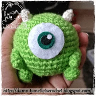 Una Mano Sostiene Un Peluche De Crochet Que Se Parece a Mike Wazowsky Con Su Tono Verde Característico Un Ojo Grande Y Dos Cuernos Pequeños El Borde Inferior Tiene Una Marca De Agua Con El Sitio Web damnitjanetletscrochetblogspotcom