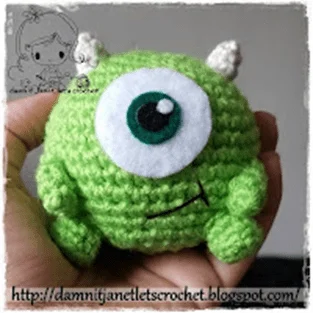 Una Mano Sostiene Una Pequeña Criatura Verde Tejida a Crochet Con Un Ojo Grande Y Dos Pequeños Cuernos Blancos Que Recuerda a Mike Wazowski La Criatura Tiene Una Sonrisa Ligeramente Traviesa El Fondo Incluye Una Ilustración De Marca De Agua Y Un Texto Que Dice damnitjanetletscrochetblogspotcom