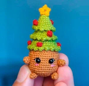 Un pequeño juguete de peluche tejido a mano que se asemeja a un árbol de navidad, con una copa verde adornada con adornos rojos y una estrella amarilla, y una base redonda marrón con lindos ojos negros y una cara sonriente. El juguete se sostiene entre dos dedos sobre un fondo azul.