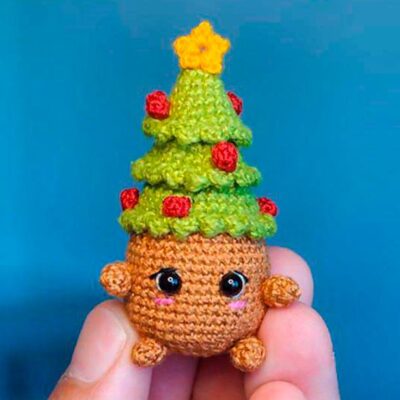 Un Pequeño Juguete De Peluche Tejido a Mano Que Se Asemeja a Un árbol De Navidad Con Una Copa Verde Adornada Con Adornos Rojos Y Una Estrella Amarilla Y Una Base Redonda Marrón Con Lindos Ojos Negros Y Una Cara Sonriente El Juguete Se Sostiene Entre Dos Dedos Sobre Un Fondo Azul