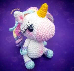 Un peluche de unicornio tejido a crochet se posa sobre un fondo morado adornado con formas de corazones tenues. El unicornio tiene un cuerno amarillo, ojos negros con pestañas y una melena y cola de color pastel. Su cuerpo es principalmente blanco y rosa con detalles en azul y morado en sus pezuñas.