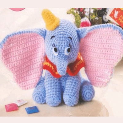 Un elefante azul tejido a crochet con grandes orejas rosadas, un sombrero amarillo y un collar rojo se sienta sobre una superficie. Delante del elefante hay pequeñas fichas de colores, una rosa y otra azul. Al fondo se ve un libro y parte de una flor.