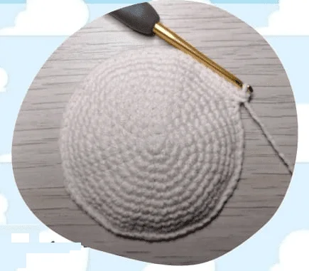 Se Muestra Un Primer Plano De Una Pieza Circular De Tejido De Crochet Beige En Proceso Se Inserta En La Tela Un Ganchillo Con Empuñadura Negra Y Punta Dorada Con Un Hilo De Hilo Blanco Adherido El Fondo Amaderado Presenta Una Superficie De Madera Y Un Diseño De Nubes Estilo Dibujos Animados