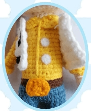 Un Primer Plano De Un Muñeco De Crochet Que Se Parece a Woody Vestido Con Una Camisa Amarilla Con Botones Blancos Pantalones Azules Y Un Cinturón Marrón Con Una Hebilla Amarilla La Muñeca También Luce Un Chaleco Blanco Con Un Estampado Negro La Imagen Está Enmarcada Por Un óvalo De Color Azul Claro Con Un Fondo De Nubes Blancas