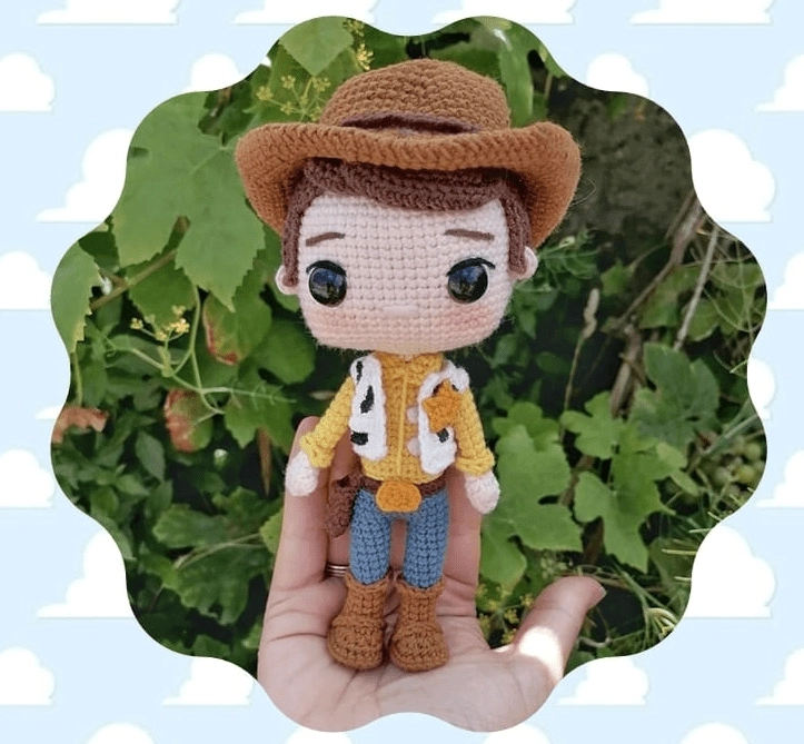 Una Mano Sostiene Un Muñeco De Crochet Vestido Como Un Vaquero Que Recuerda a Woody Con Un Sombrero Marrón Camisa Amarilla Pantalón Azul Y Botas Marrones El Fondo Presenta Follaje Verde Y Un Borde Azul Cielo Festoneado Con Nubes Blancas