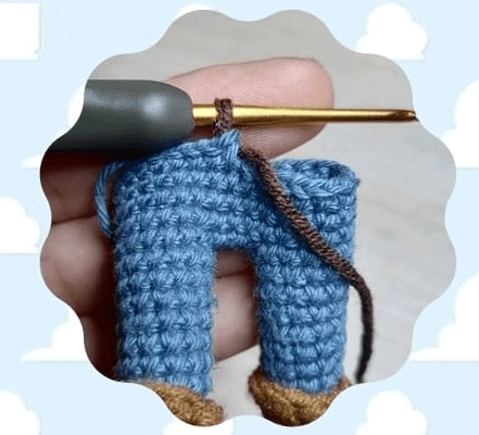 La Mano De Una Persona Sosteniendo Un Ganchillo Y Trabajando En La Confección De Un Pequeño Par De Pantalones Azules Con Zapatos Marrones El Fondo Presenta Un Patrón De Nubes Caprichoso Que Recuerda a Una Escena De Las Aventuras De Woody