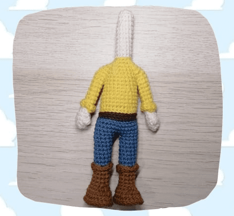 Una Figura Tejida a Crochet Con Una Cabeza Larga Y Blanca Y Sin Rasgos Faciales Vestida Con Una Camisa De Color Amarillo Brillante Pantalones Azules Un Cinturón Marrón Y Botas Marrones El Fondo Tiene Una Textura Leñosa Con Un Patrón De Cielo Nublado Alrededor De Los Bordes