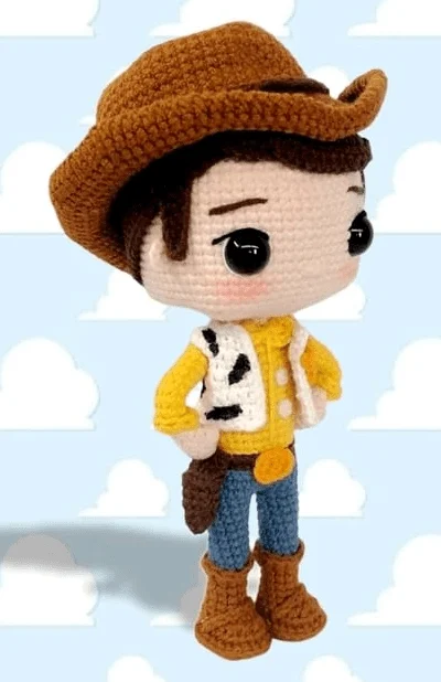 Un Muñeco De Ganchillo Que Se Parece Al Querido Personaje Woody Se Alza Sobre Un Fondo Azul Claro Con Nubes Blancas El Muñeco Lleva Un Sombrero De Vaquero Marrón Una Camisa Amarilla Un Chaleco Blanco Con Manchas Negras Pantalones Azules Y Botas Marrones Capturando Un Diseño Divertido Y Detallado