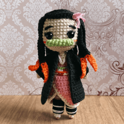 Una muñeca de ganchillo presenta un diseño complejo con cabello largo y negro con reflejos rojos, un vestido rosa, un abrigo negro y un accesorio para el cabello rosa. La muñeca tiene una boca verde que recuerda a Nezuko y se alza sobre un fondo estampado, mostrando fina artesanía y atención al detalle.