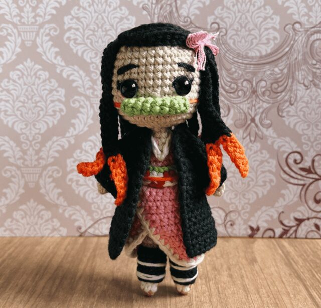 Una Muñeca De Ganchillo Presenta Un Diseño Complejo Con Cabello Largo Y Negro Con Reflejos Rojos Un Vestido Rosa Un Abrigo Negro Y Un Accesorio Para El Cabello Rosa La Muñeca Tiene Una Boca Verde Que Recuerda a Nezuko Y Se Alza Sobre Un Fondo Estampado Mostrando Fina Artesanía Y Atención Al Detalle