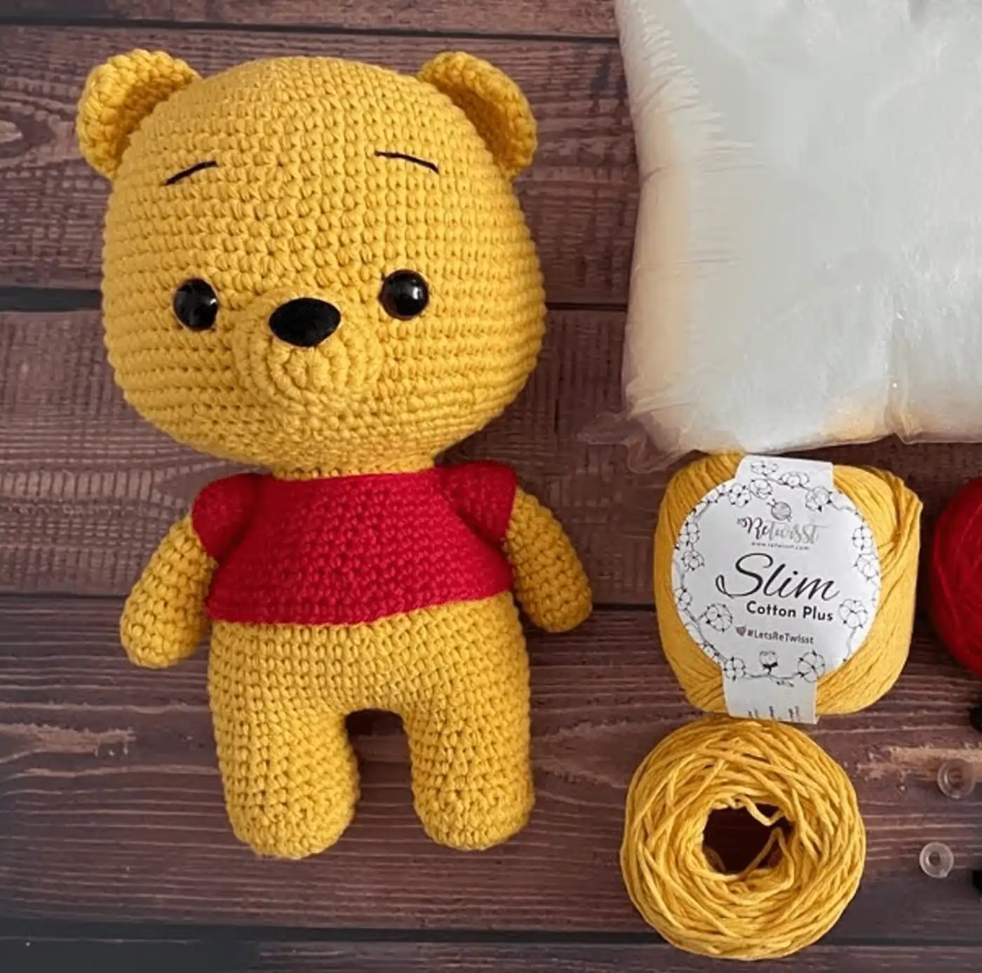 Un muñeco Winnie the Pooh amigurumi terminado, tejido a ganchillo, sentado junto a un ovillo de lana color mostaza.