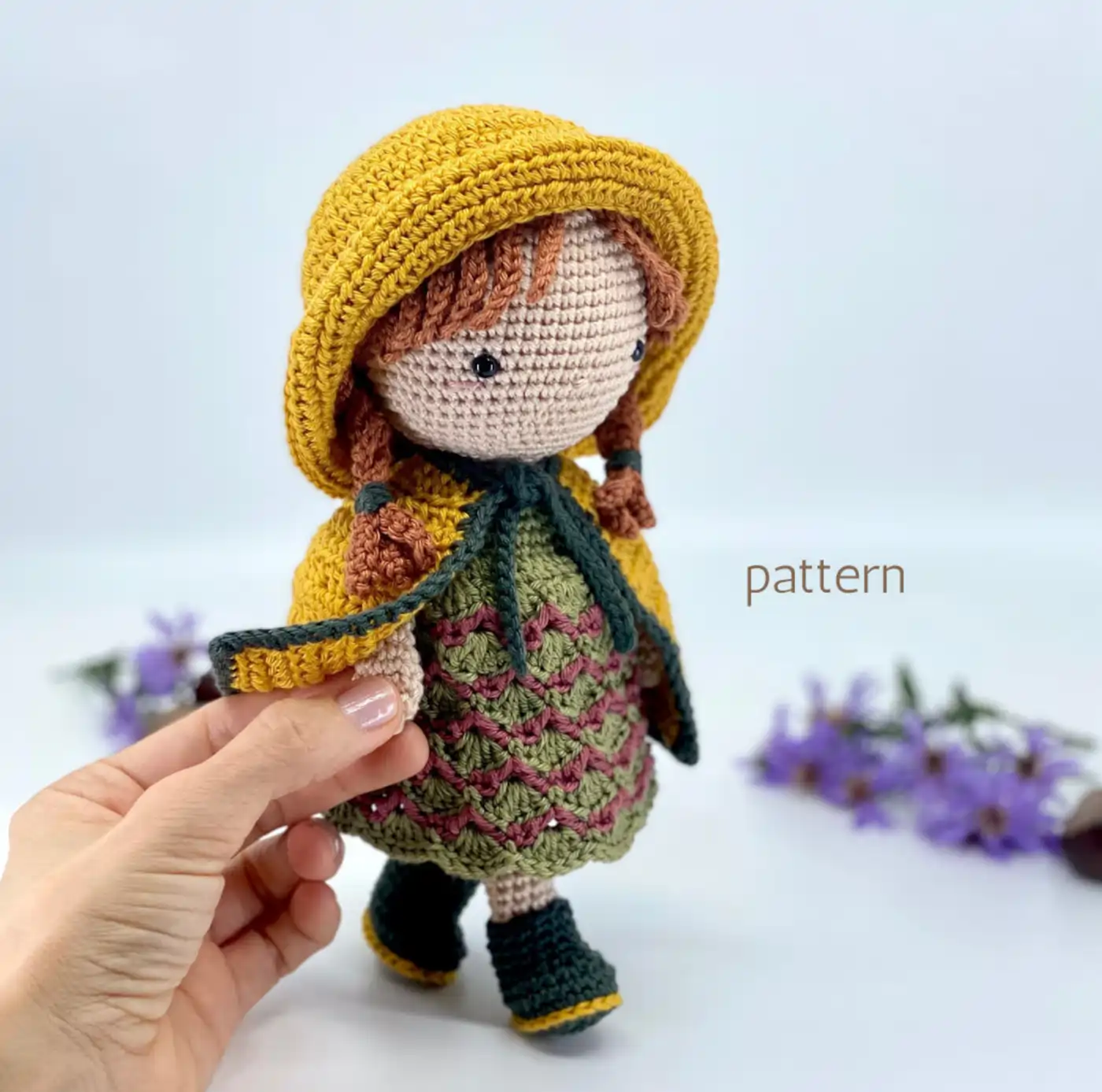 Muñeca amigurumi Baby Alice terminada, con vestido verde, sombrero rojo y trenzas rubias, sentada sobre una superficie de madera.