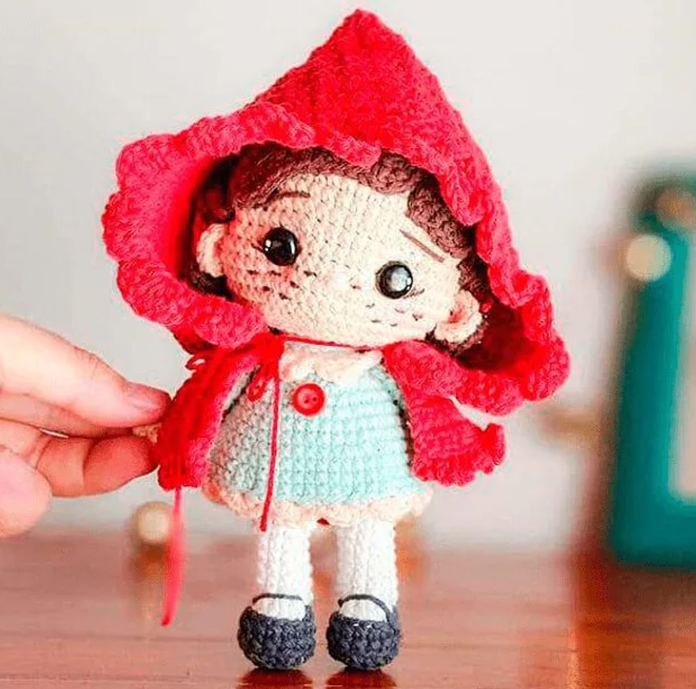 Muñeco tejido de Caperucita Roja amigurumi con su capa roja y vestido verde, terminada sobre una superficie de madera.