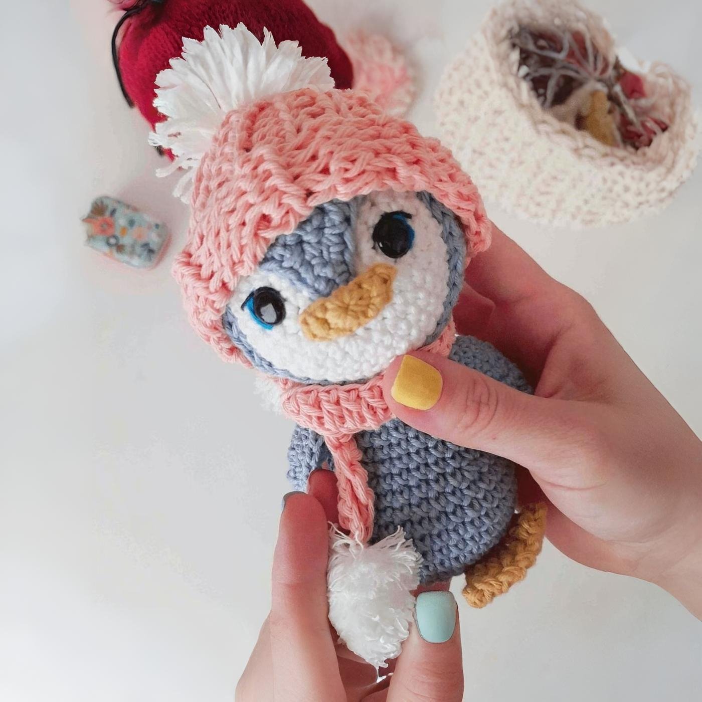 Pingüino amigurumi tejido a crochet de color gris con gorro y bufanda rosa sobre fondo claro