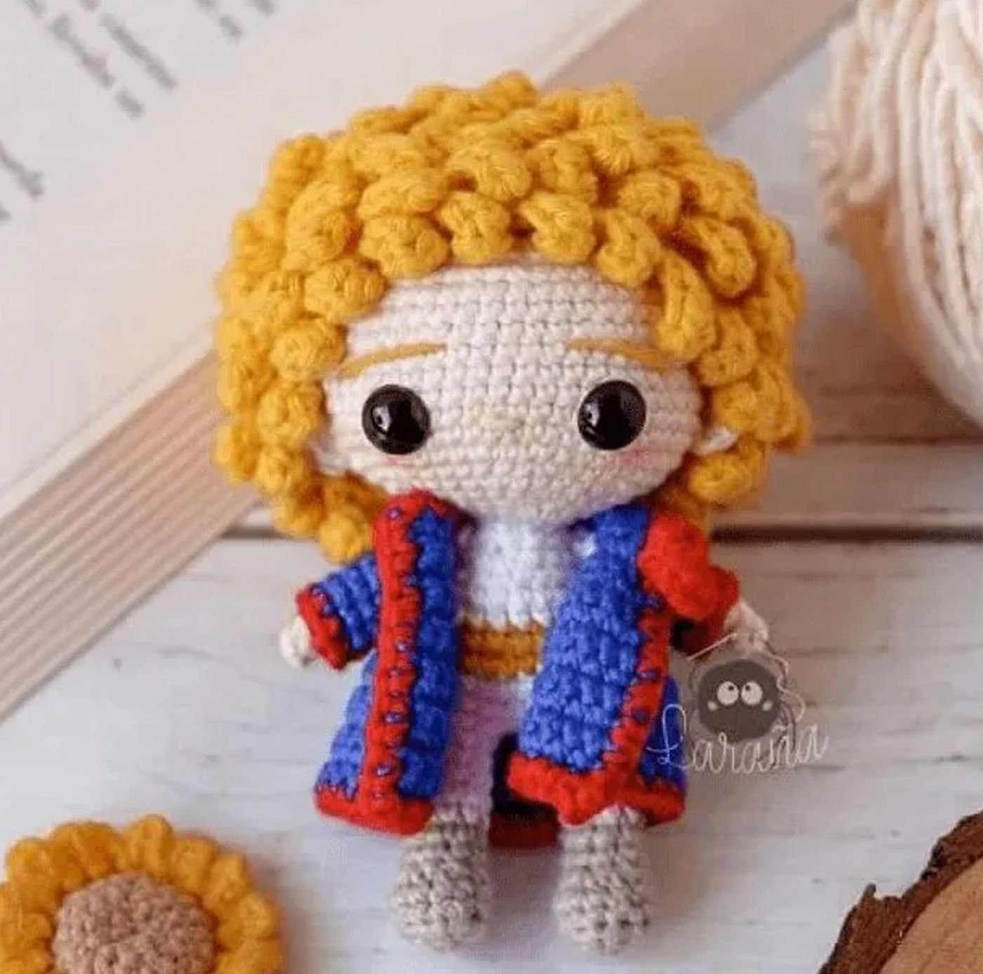 Muñeco tejido del Principito amigurumi terminado, con su traje azul, cabello rubio y una rosa roja en la mano.