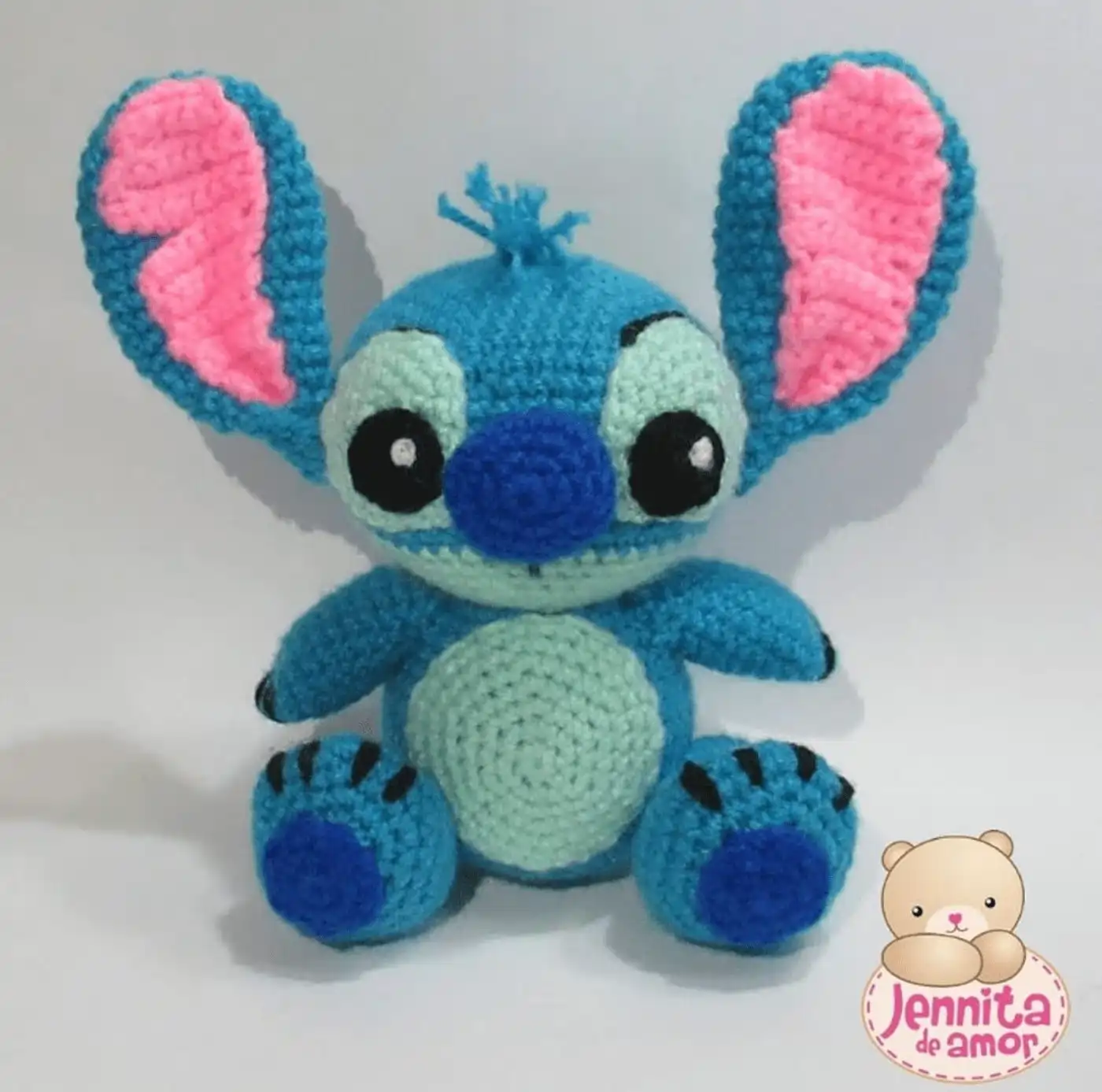 Muñeco tejido de Stitch amigurumi color azul con orejas grandes y detalles en celeste, sentado sobre una mesa de madera.